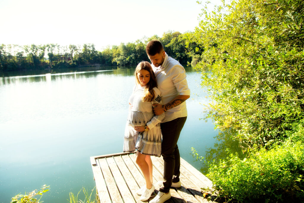 Ein Paar steht auf einem Holzsteg am See in Wiesbaden. Die Frau hält ihren Babybauch, der Partner legt seine Hände an ihre Oberarme und blickt auf den Bauch. Goldenes Sonnenlicht fällt seitlich ins Bild.