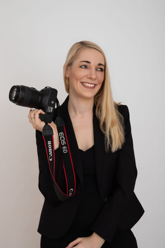 Hochzeitsfotografin aus Mainz sitzend in schwarzer Kleidung und blickt konzentriert auf ihre Kamera, die sie in den Händen hält. Natürliches Businessportrait vor hellem Hintergrund.