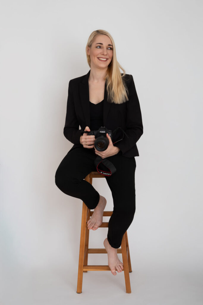 Hochzeitsfotografin aus Mainz sitzt auf einem Hocker, hält ihre Kamera fest in den Händen und blickt seitlich aus dem Bild. Authentisches Businessportrait in schwarzer Kleidung.