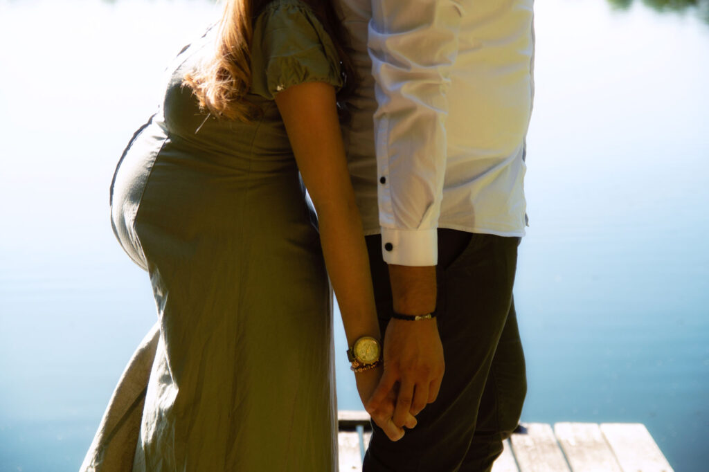 Ein Paar steht am See Rücken an Rücken und hält sich an den Händen. Die Gesichter sind nicht sichtbar, der Fokus liegt auf dem Babybauch der werdenden Mutter.
